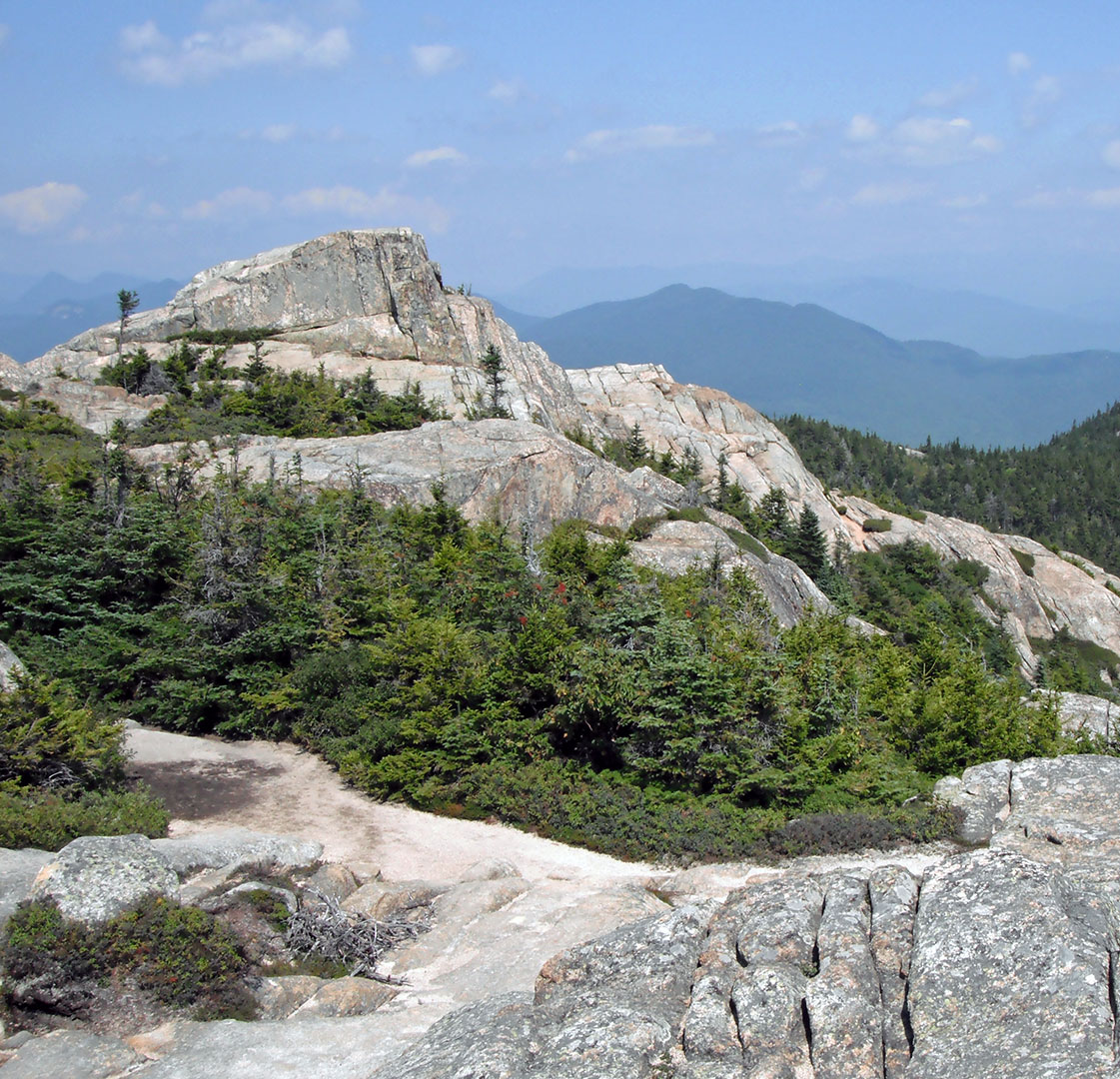 Rocky mountain summit with evergreen trees and a dirt trail under a blue sky with distant hazy mountains.
